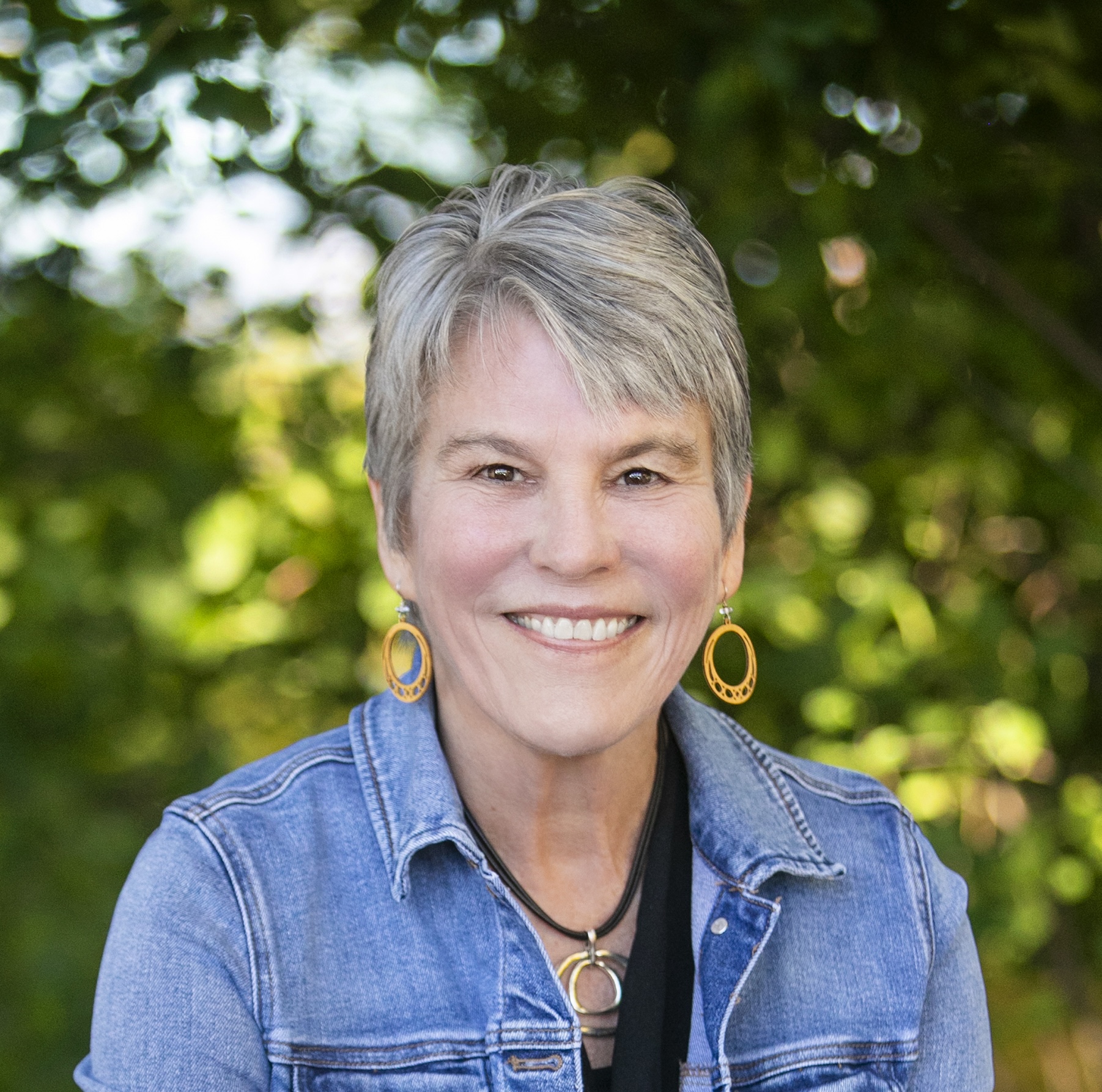Jill Hughes of New Roots counseling is pictured in an outdoor portrait with a vibrant green background. She has short grey hair and wears a black top and denim jacket.